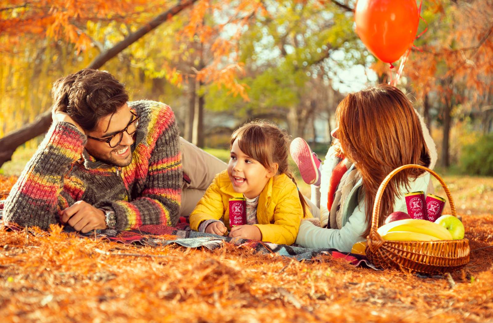 family of mother father and daughter laying in grass autumn field with happy belly kombucha apple spice cans in picnic basket banner