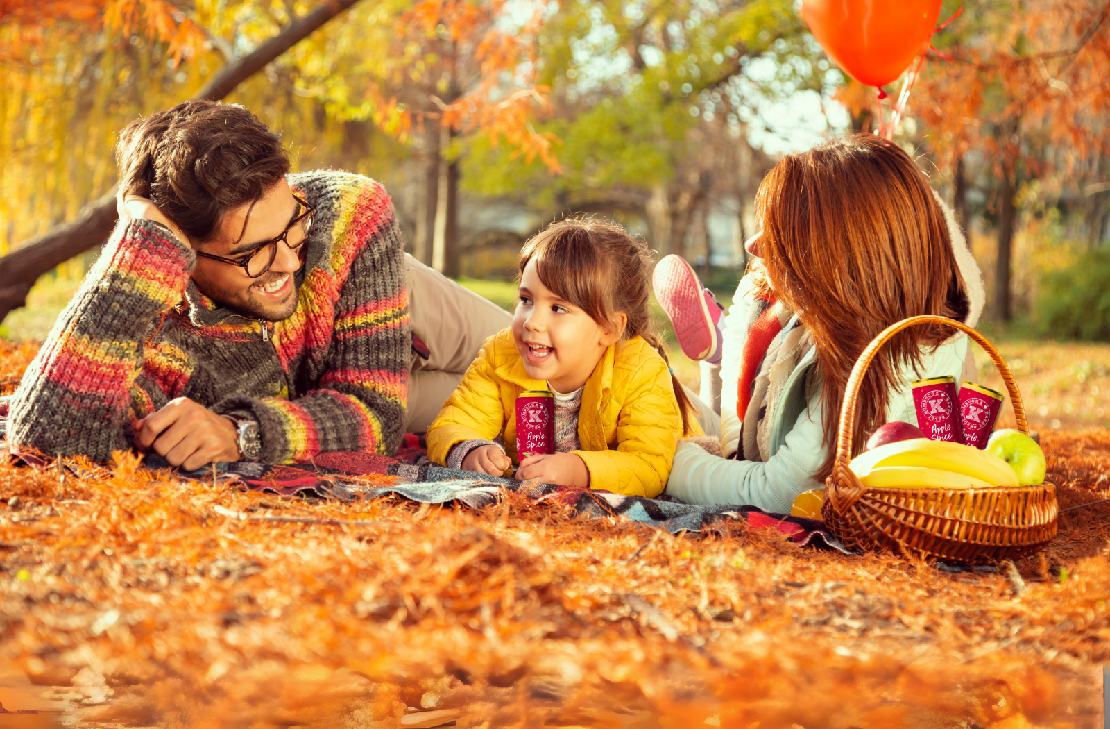 family of mother father and daughter laying in grass autumn field with happy belly kombucha apple spice cans in picnic basket banner