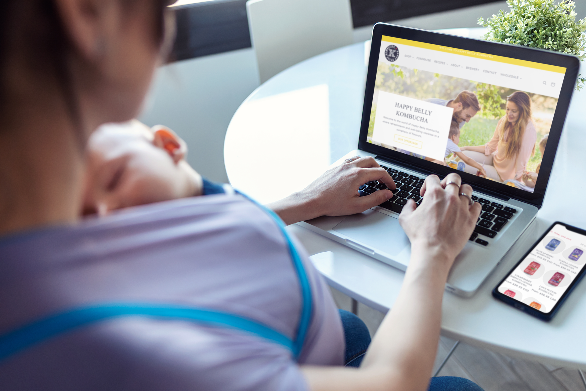 mother with baby on chest browsing happy belly kombucha website shop in her laptop and phone